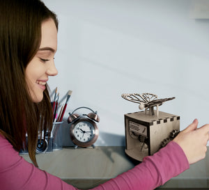 Model shown smiling while cranking wheel of automaton. The automaton is placed on desk.